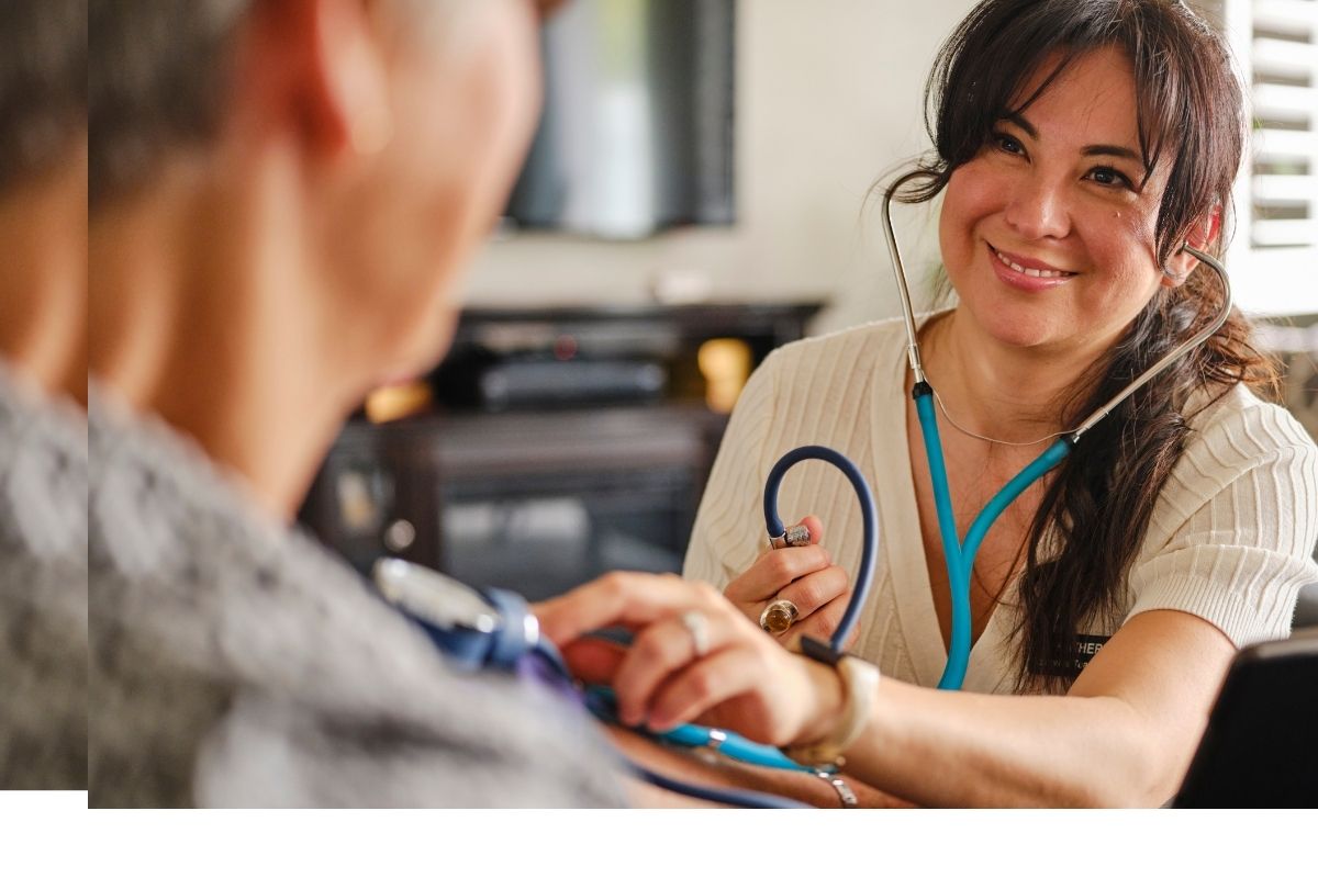 Healthcare worker smiling while taking an older adult’s blood pressure at home with a stethoscope