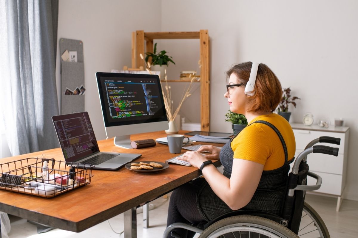 Woman using a wheelchair working at a desk with headphones, coding on dual monitors in a home office