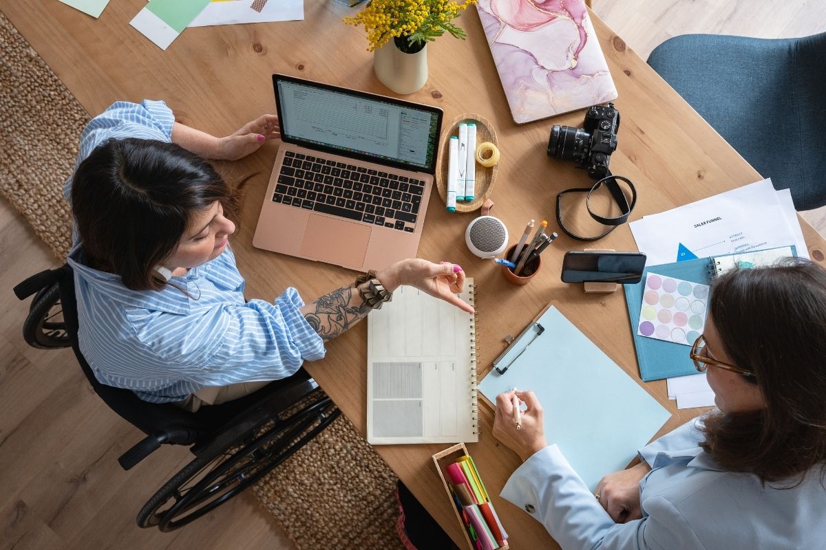 Overhead view of two colleagues working at a desk, one using a wheelchair, with a laptop, papers and a camera on the table