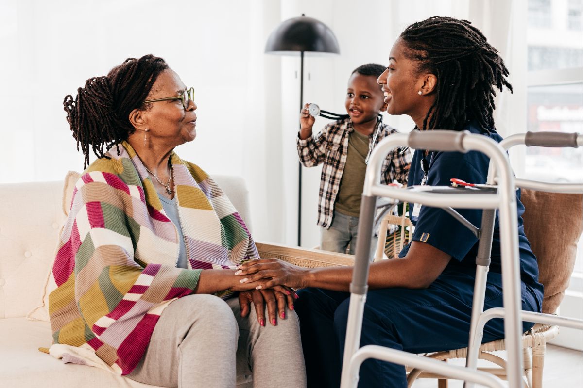 Nurse talking with an older woman at home, a walker beside them and a child in the background