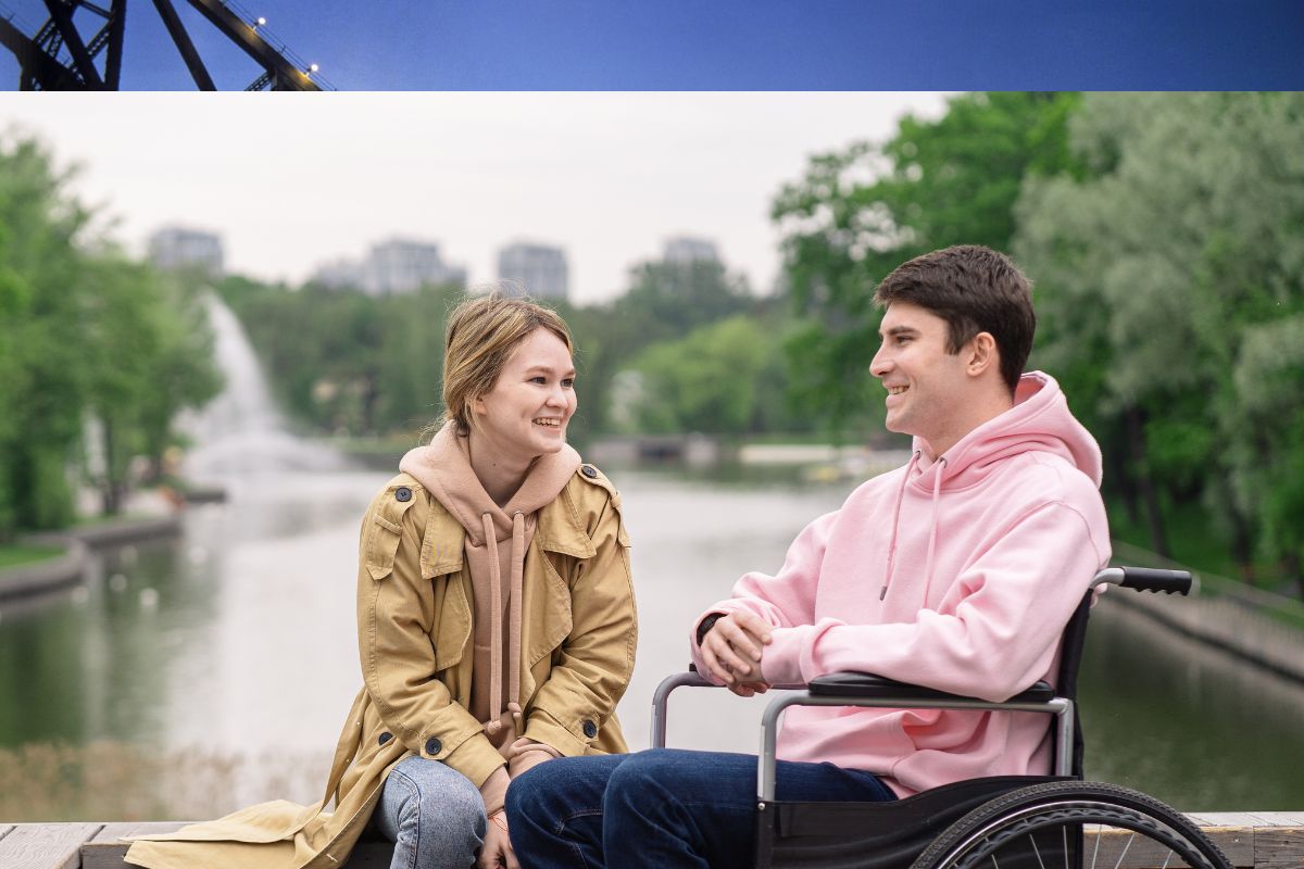 Two friends chatting by a riverside; one person uses a wheelchair, both smiling on a wooden deck.