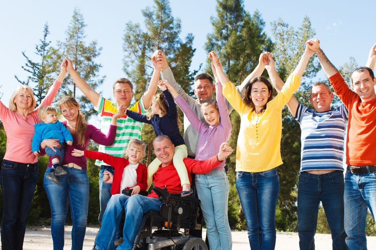 Group of families outdoors holding hands up, including a man using a wheelchair, smiling in front of trees
