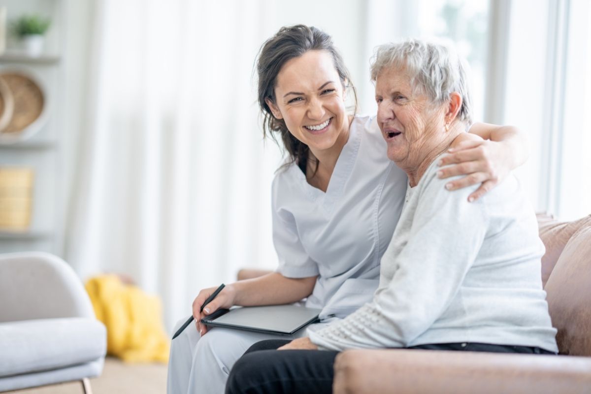 Support worker with a clipboard smiling and hugging an older woman on a couch